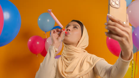 Studioporträt-Einer-Frau,-Die-Ein-Selfie-Mit-Hijab-Macht-Und-Geburtstag-Mit-Partygebläse-Feiert,-Umgeben-Von-Luftballons